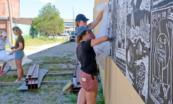 Artists add printed paper to a wall in a rural setting with old building in the background.