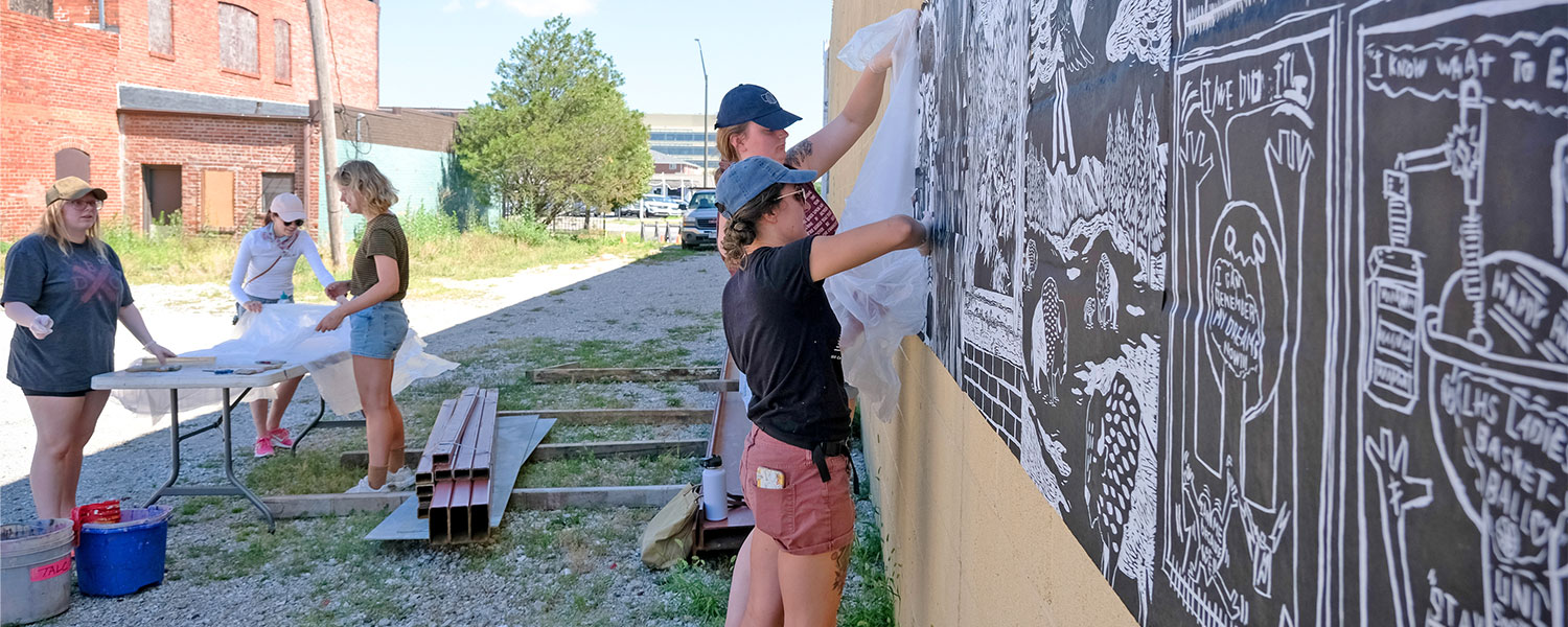 Artists add printed paper to a wall in a rural setting with old building in the background.