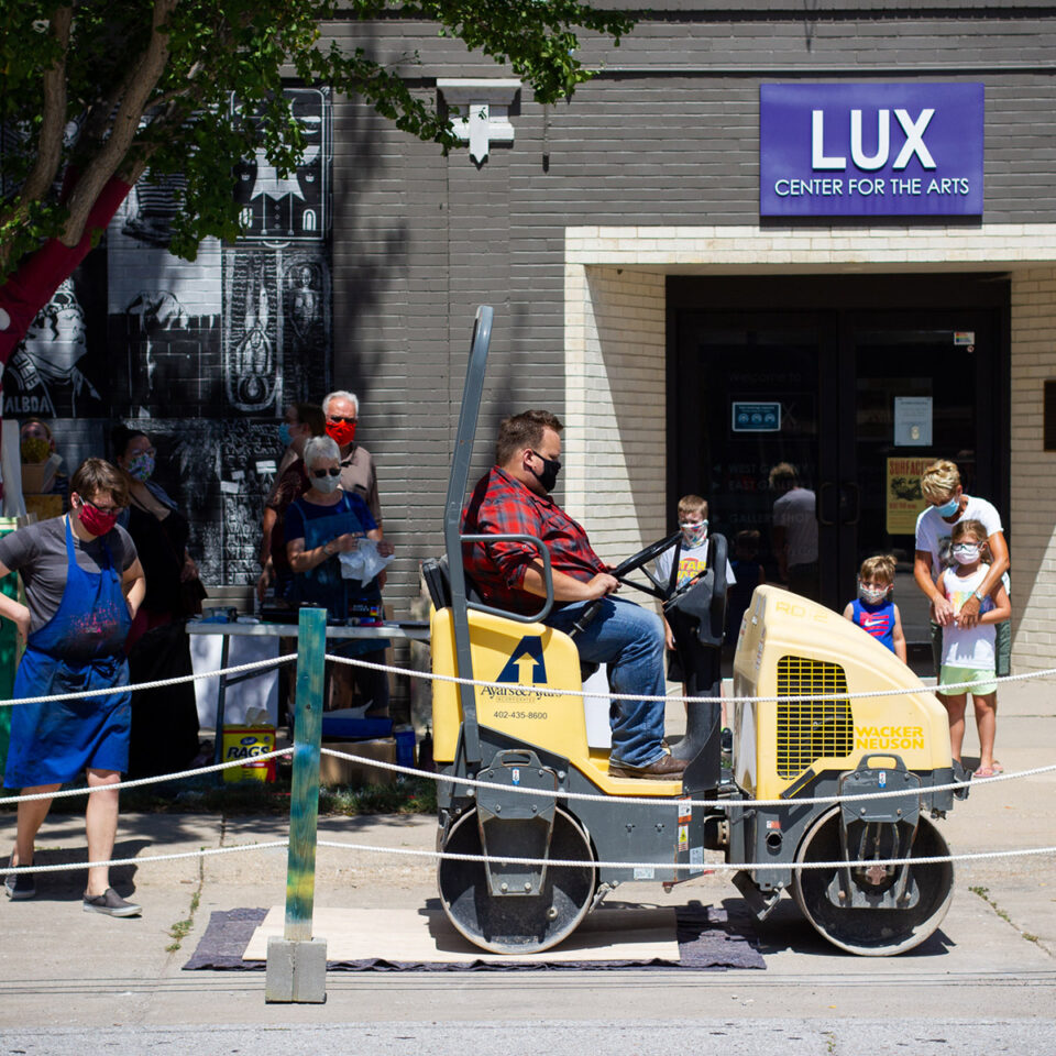 A man rides a steamroller over a paper and carving for a relief print technique in front of the Lux Center for the Arts in Nebraska with an audience of onlookers. 