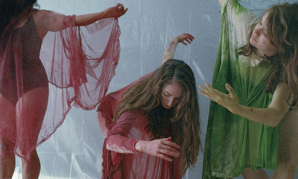 Three light-skinned female presenting individuals wear leotards and sheer draped fabric in poses that appear symbolic and dramatic. The people in the middle and left have burgundy fabrics and on the right has green. There is a white backdrop.