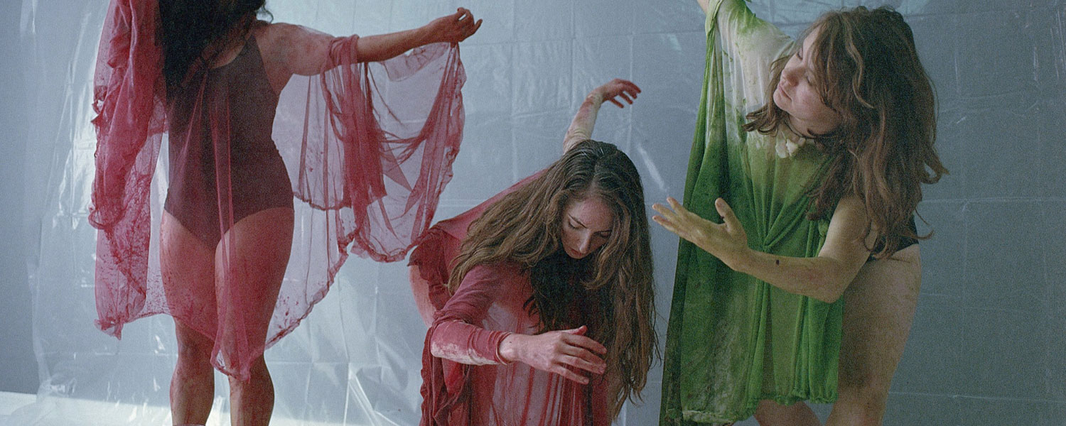 Three light-skinned female presenting individuals wear leotards and sheer draped fabric in poses that appear symbolic and dramatic. The people in the middle and left have burgundy fabrics and on the right has green. There is a white backdrop.