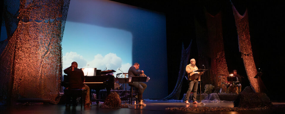 Man in tan jacket talks into a microphone with his arm raised on a large state. A projection in the background shows a blue sky and clouds as if taken from above the clouds in an airplane. Musicians including a piano at left surround the man.