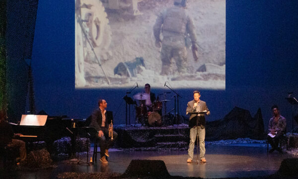 Man stands at a podium reading a story holding a microphone on a stage with a projection behind him of a man in armed combat. A musician to his right hold a trumpet, and a group of musicians to his left are sitting at drums, a base guitar, and percussion. There is dramatic lighting with green up lights and a large stage.