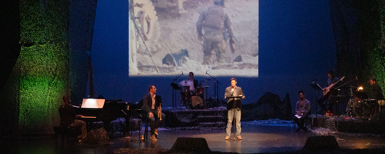 Man stands at a podium reading a story holding a microphone on a stage with a projection behind him of a man in armed combat. A musician to his right hold a trumpet, and a group of musicians to his left are sitting at drums, a base guitar, and percussion. There is dramatic lighting with green up lights and a large stage.