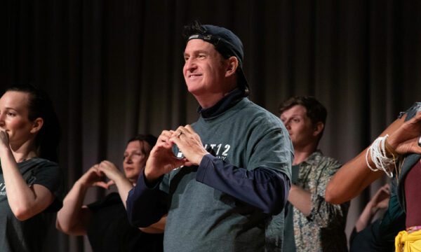 A group of people standing looking off to the left while holding their hands at their chest, forming a shape of a heart.