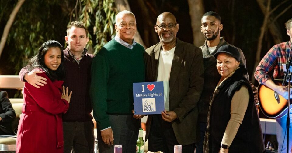 A group of six people smiling while standing in front of a small table with food and drinks on it. The person in the center is holding a sign that says "I love Military Nights at La Jolla Playhouse."