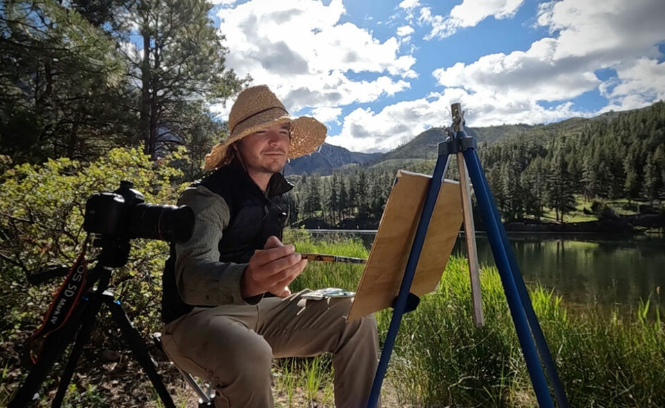 Man wearing a large straw hat paints on an easel in a beautiful outdoor wilderness setting next to a river with a camera set up next to him.