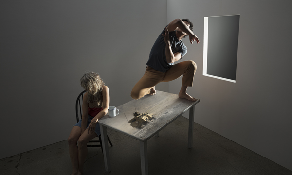 Dancers Felip Barrueto and Marit Brook-Kothlow are in a room with one window, a chair, a table, and a mug. Marit sits on the chair with her head down as Felipe hits a crouched pose on the table with their elbow up over their face. The table has an image of an army tank projected on it.
