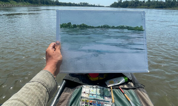 A man holds up a watercolor painting from a canoe in the middle of a river.
