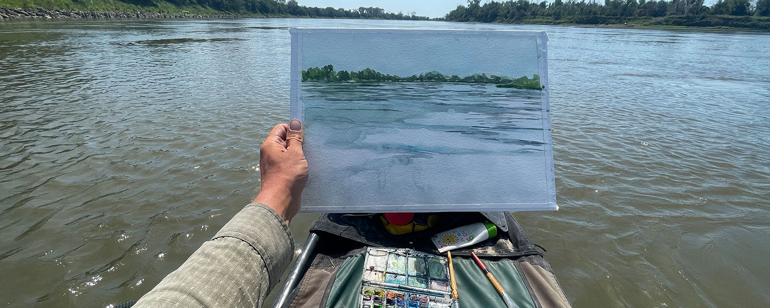 A man holds up a watercolor painting from a canoe in the middle of a river.