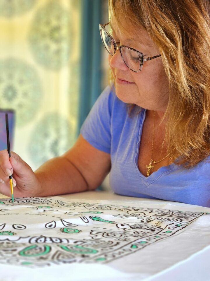 A woman with long light brown hair, wearing glasses, is seated while holding a paintbrush and looking down at a cloth canvas.