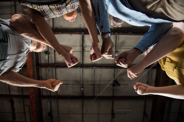 Image looking up of a group of people with their arms extended and hands clenched in a fist. Their arms are the focus of the image. Overhead is a ceiling with lighting tresses.
