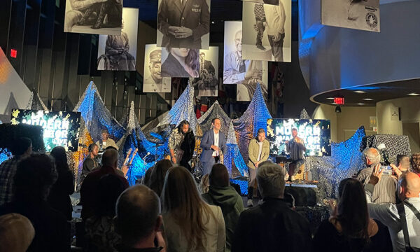 Posters of veterans hang from the ceiling of an interior curved room with a stage and densely seated audience. There is blue and white lighting illuminating three male-presenting individuals onstage.