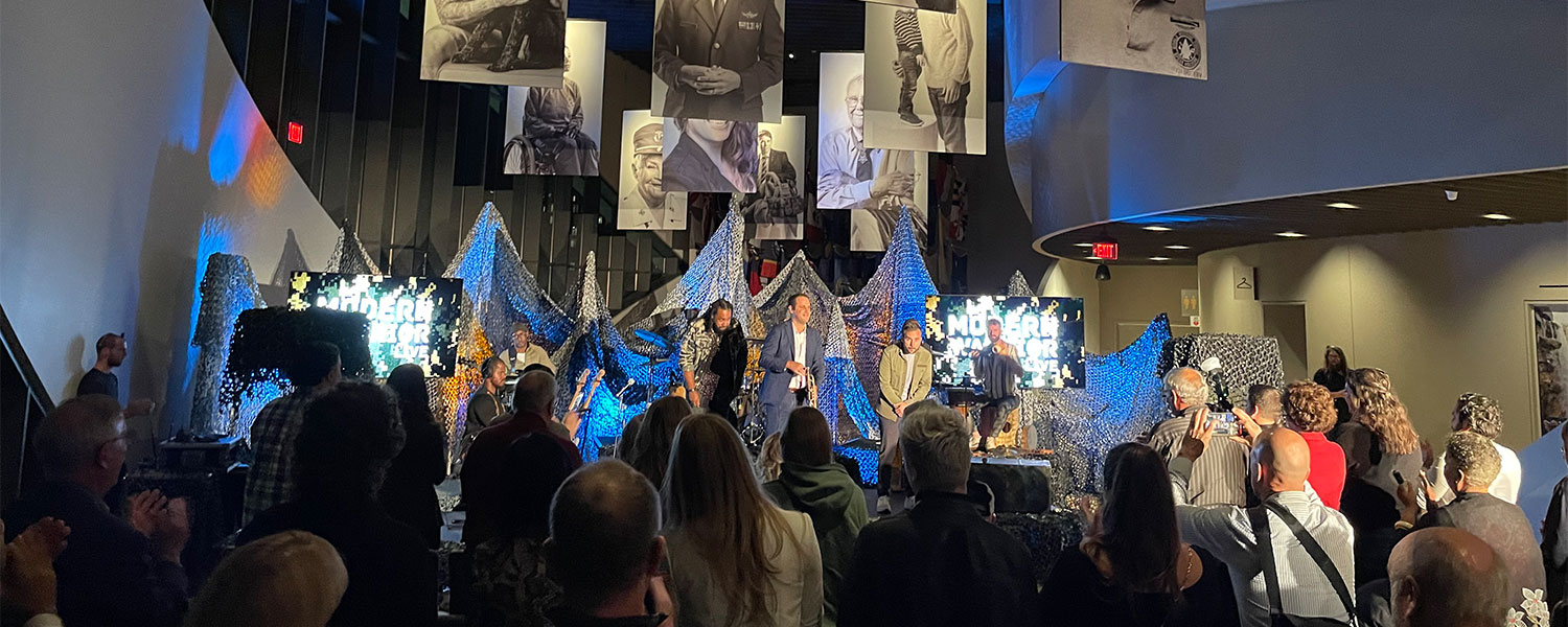 Posters of veterans hang from the ceiling of an interior curved room with a stage and densely seated audience. There is blue and white lighting illuminating three male-presenting individuals onstage.