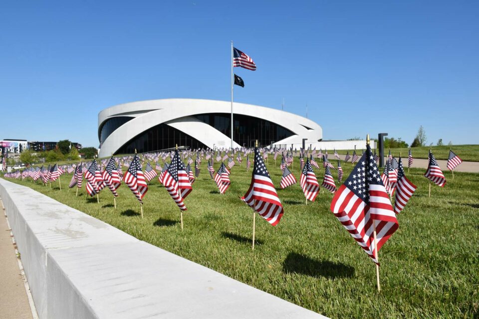 A curved building with wrapped wall structures sits low in a landscape of clear blue sky and green grass with USA flags dotting the lawn.