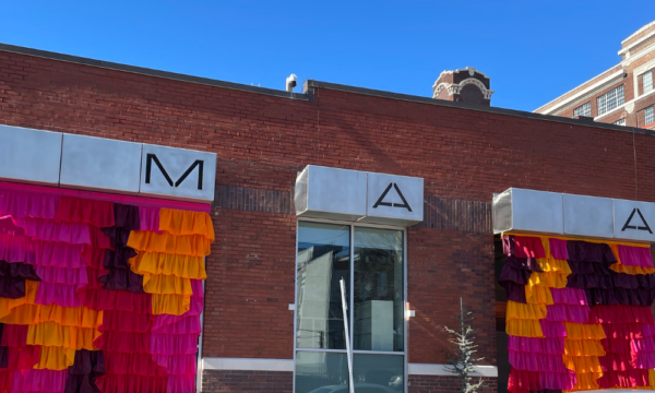 M-AAA's brick building exterior with a large bright pink, purple, and orange fabric installation on the two large front windows.