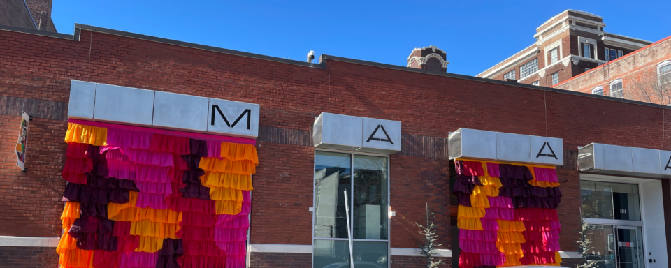 M-AAA's brick building exterior with a large bright pink, purple, and orange fabric installation on the two large front windows.