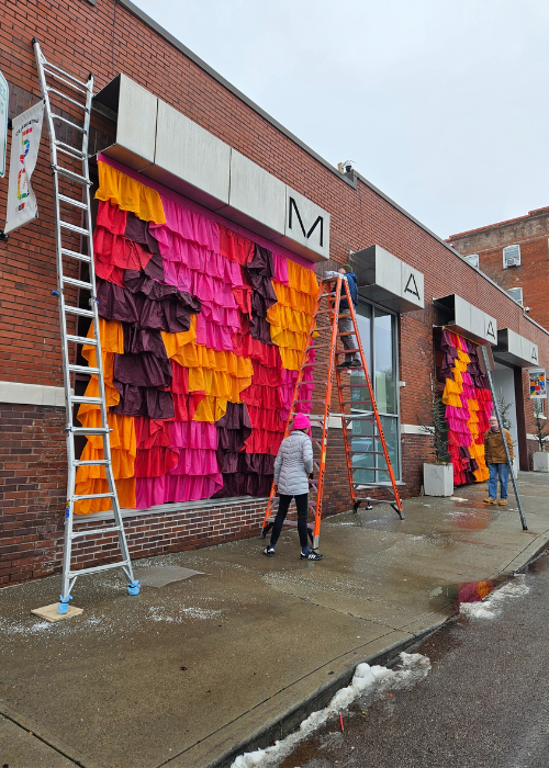 Exterior photograph of a brick gallery building and bright pink, purple, and orange fabrics hanging the entire height of the building. There are two large ladders and two people installing the work with a cold, gray looking sky and wet ground.