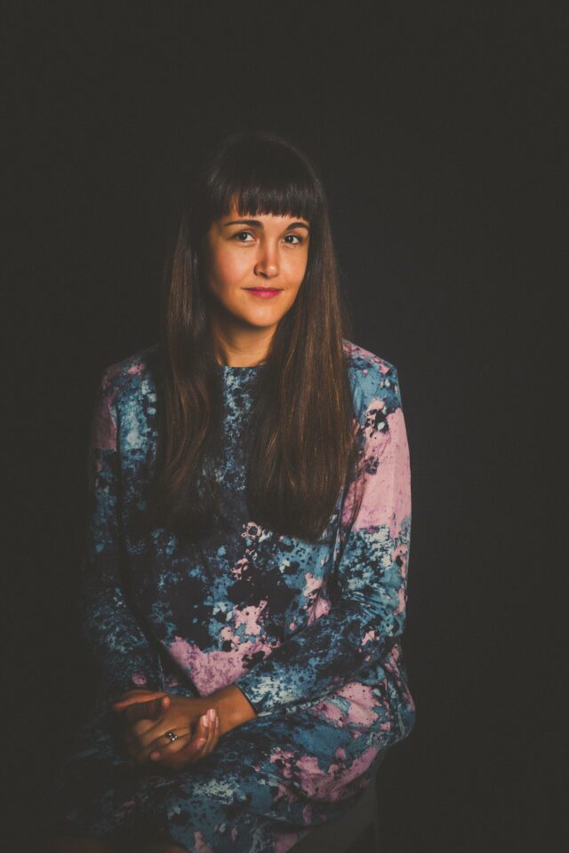 Female-presenting individual sitting for a posed portrait with long dark hair, bangs, and wearing a blue and purple dress.