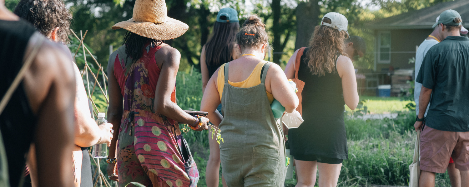 A group of people appearing to be a variety of ages, ethnicities, and genders, walk together outdoors on a farm in the sunshine while picking herbs and flowers.
