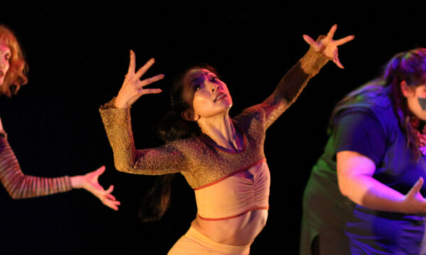 Three female-presenting individuals dance with arms in dynamic shapes reaching up and out. The person on the left wears red dancewear, in the middle wears tan, and on the right wears blue. The background is black and lighting is very dark and dramatic.