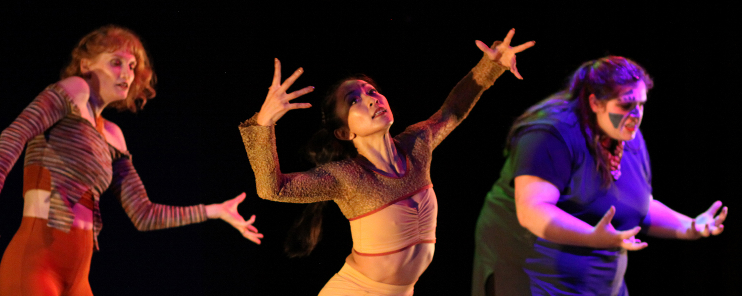 Three female-presenting individuals dance with arms in dynamic shapes reaching up and out. The person on the left wears red dancewear, in the middle wears tan, and on the right wears blue. The background is black and lighting is very dark and dramatic.