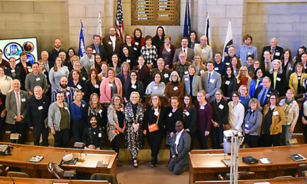 A large group of arts advocates stand together with legislators in a historical building with wooden seating, state and national flags and stone walls behind them.
