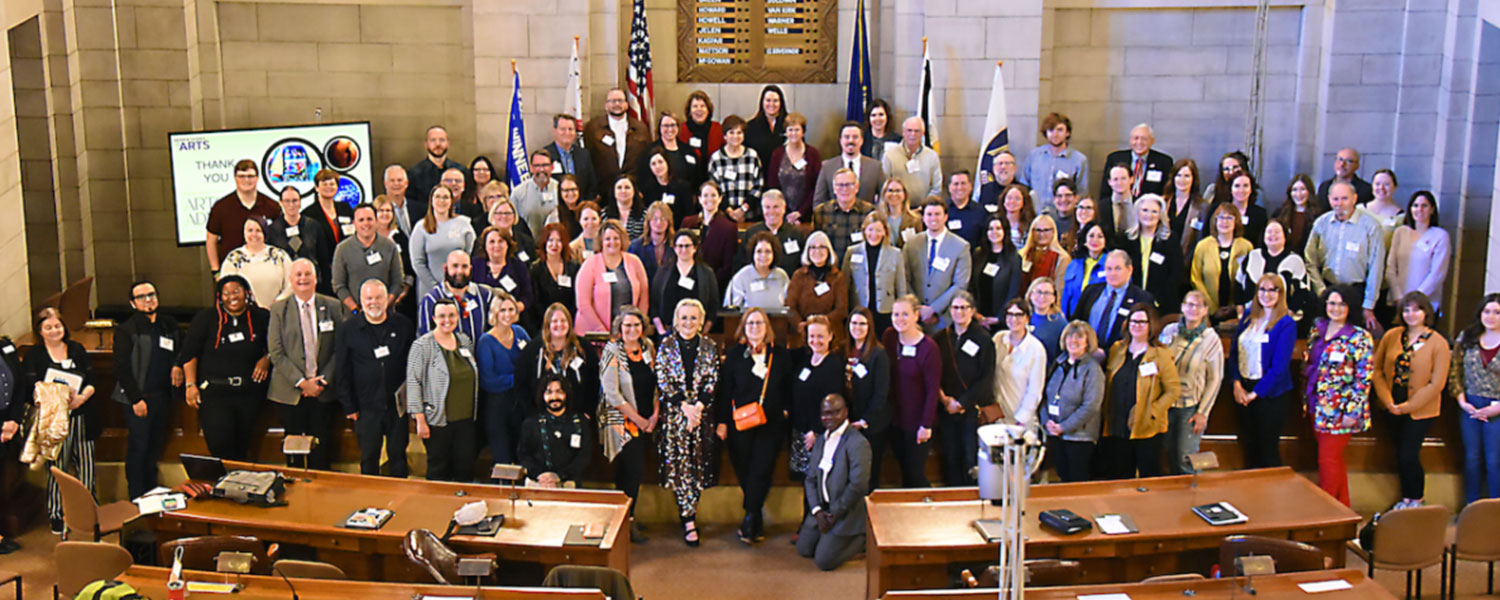 A large group of arts advocates stand together with legislators in a historical building with wooden seating, state and national flags and stone walls behind them.