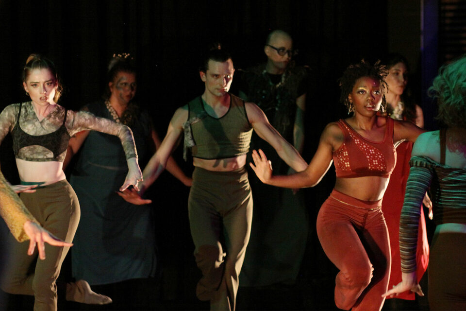 Three dancers in dramatic lighting and leotards with pants appear to march towards camera. They are surrounded by vocalists, slightly blurred in the background and a few dancers out of focus in the foreground. The dancers have costumes with tones of brown, greens, yellows, and oranges, with sparkly earrings and jewelry.