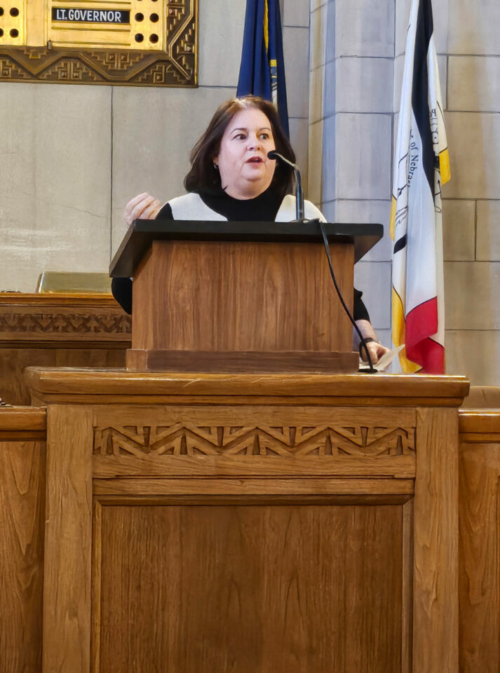 Woman with dark hair speaks into a microphone behind a decoratively-carved wooden podium with flags behind her.