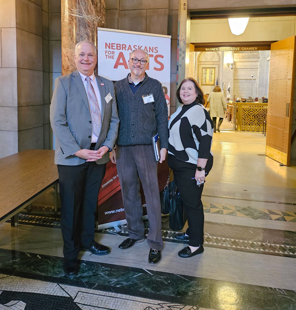 Three people dressed in business-style clothing stand on mosaic tile floor in a large interior historic building with a temporary banner sign behind them that says Nebraskans for the Arts.
