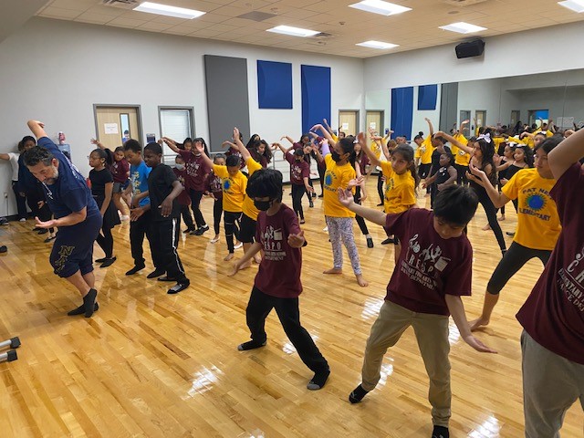 Young school children take a dance class in a school gymnasium.