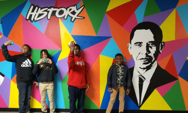 Four dark-skinned young boys stand in front of a vibrant multi-colored mural with triangular shapes of red, blue, purple, yellow, orange, prink with also paintings of Frida Kahlo and Barak Obama, and a text that says 