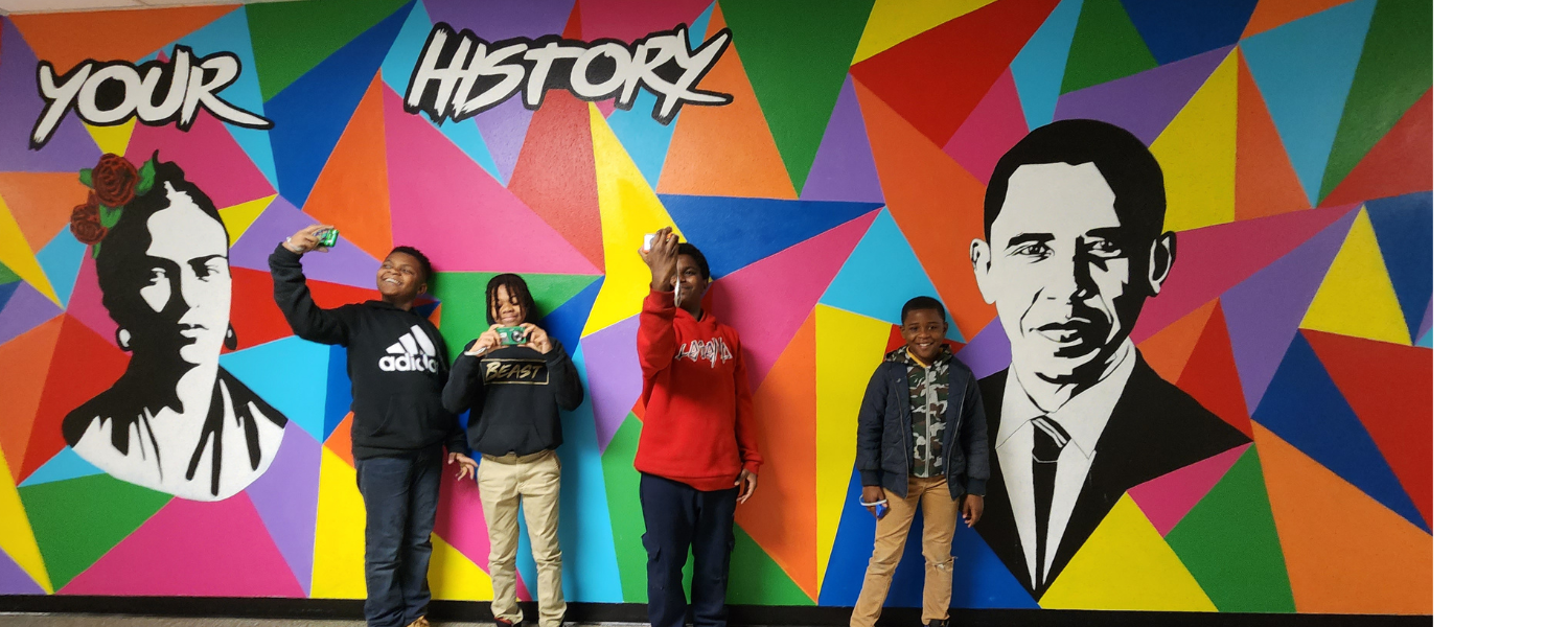 Four dark-skinned young boys stand in front of a vibrant multi-colored mural with triangular shapes of red, blue, purple, yellow, orange, prink with also paintings of Frida Kahlo and Barak Obama, and a text that says 