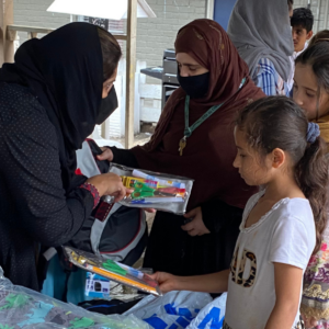 Woman wearing traditional Muslim headscarf hands bags of art supplies to children standing in a queue.