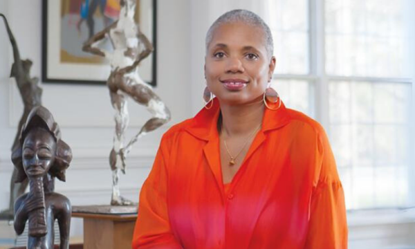 African American woman sits wearing a orange shirt with sculptures and artwork in the background.