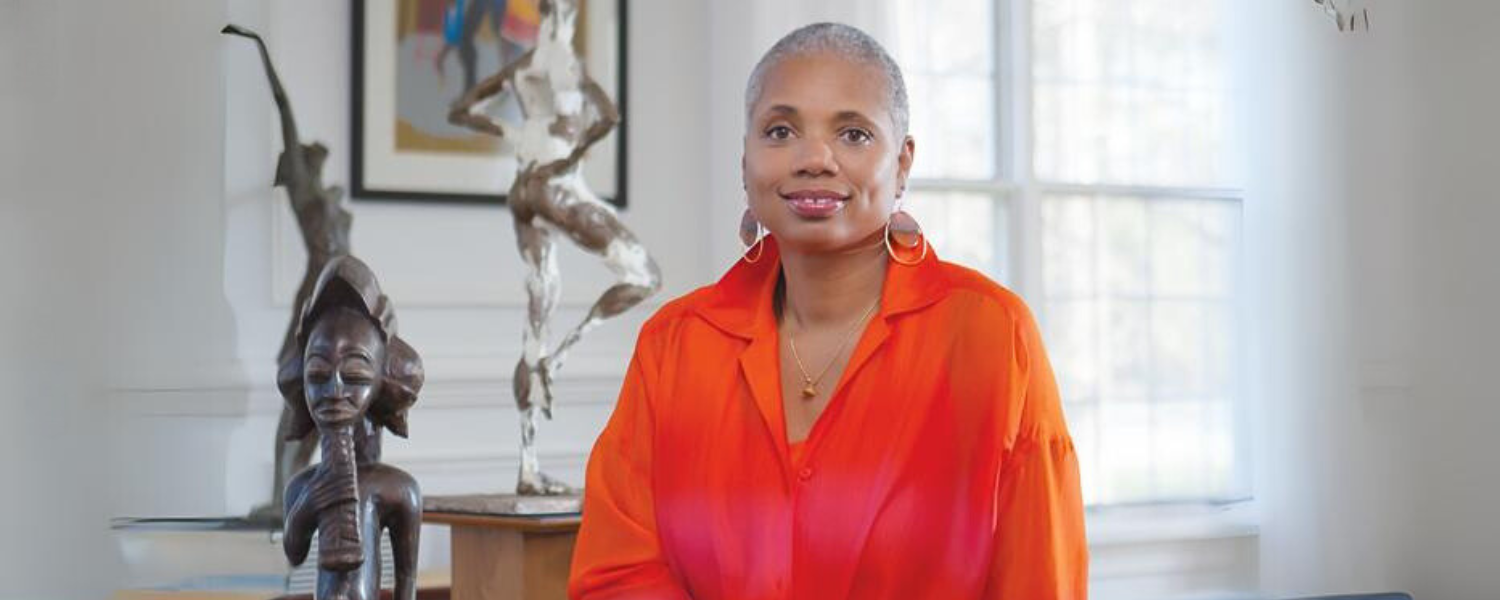 African American woman sits wearing a orange shirt with sculptures and artwork in the background.