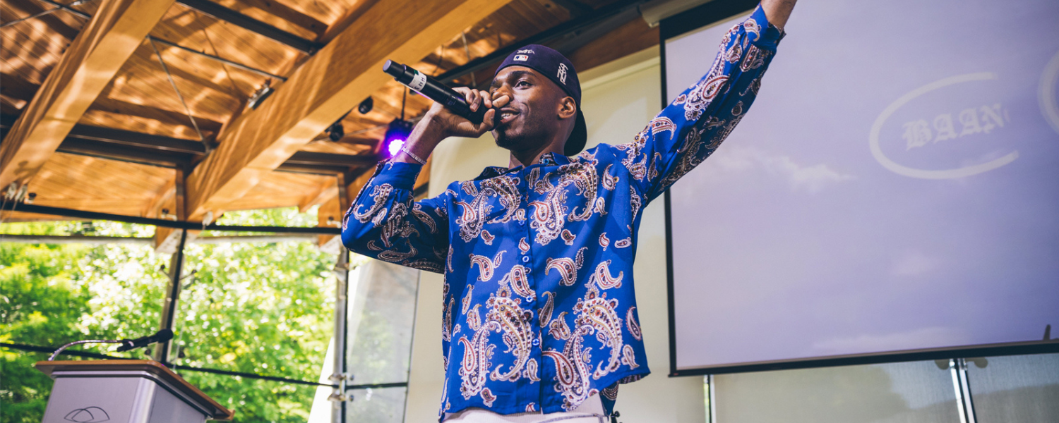 Man with blue printed shirt speaks into a microphone with his hand in air.