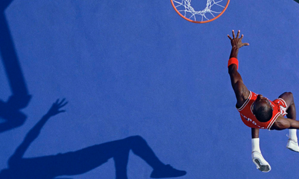 A bird's eye view action shot of professional basketball athlete Michael Jordan mid-air on the way to dunking a basketball in a nearby hoop with a royal blue court floor.