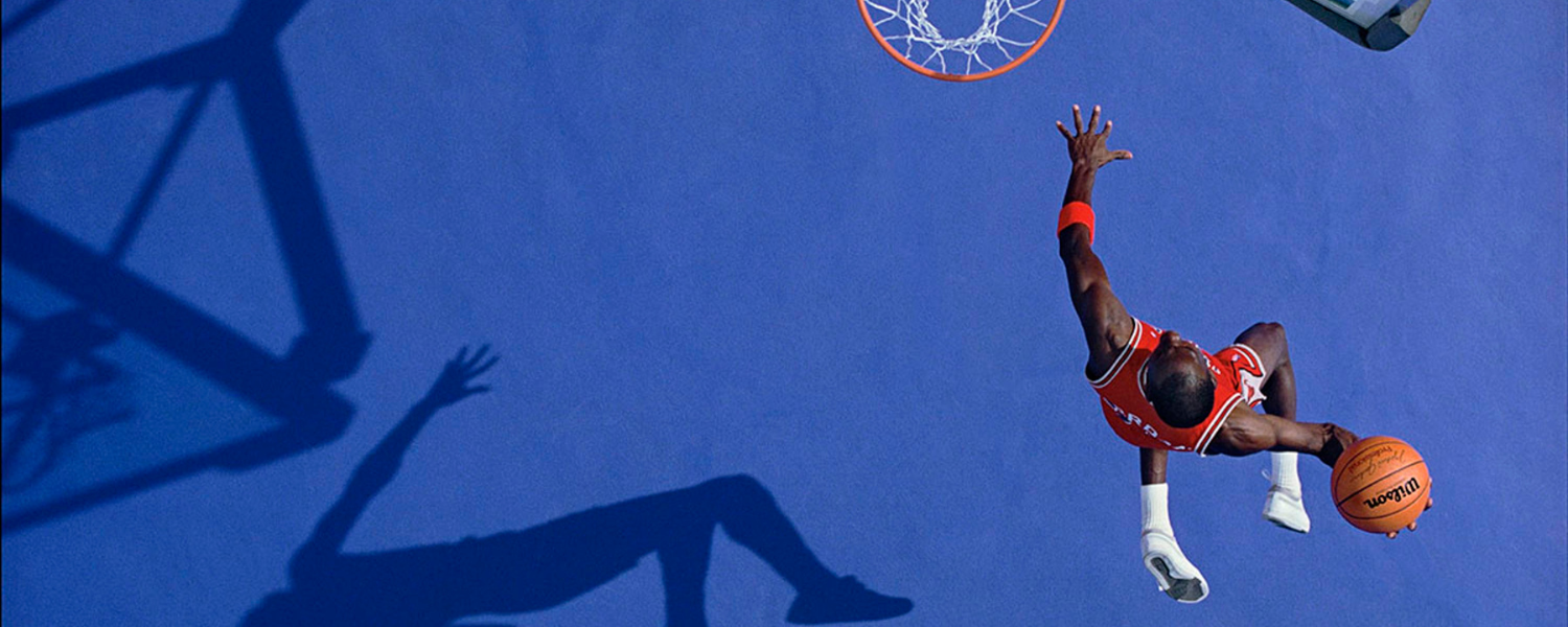 A bird's eye view action shot of professional basketball athlete Michael Jordan mid-air on the way to dunking a basketball in a nearby hoop with a royal blue court floor.