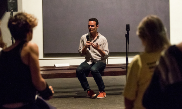 Man sits on a gallery bench with a microphone and hands expressively in motion. A large dark painting by Mark Rothko is behind him and people sit in the audience in the foreground, out of the focus of the camera.