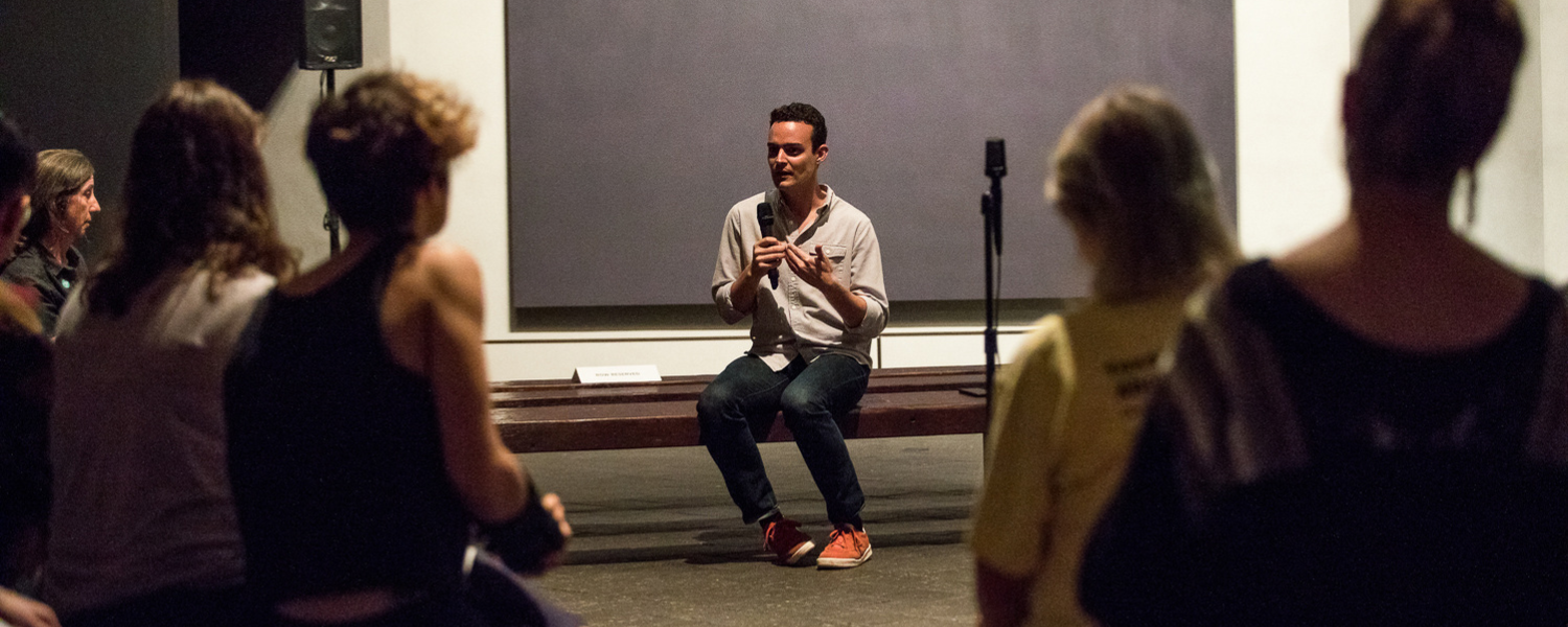 Man sits on a gallery bench with a microphone and hands expressively in motion. A large dark painting by Mark Rothko is behind him and people sit in the audience in the foreground, out of the focus of the camera.