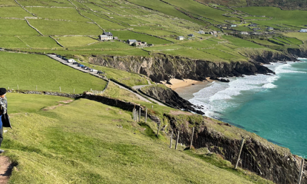 A woman wearing a coat and hat walks along a path on the top of a rocky cliff slope and looks back at a grassy green landscape and ocean below.