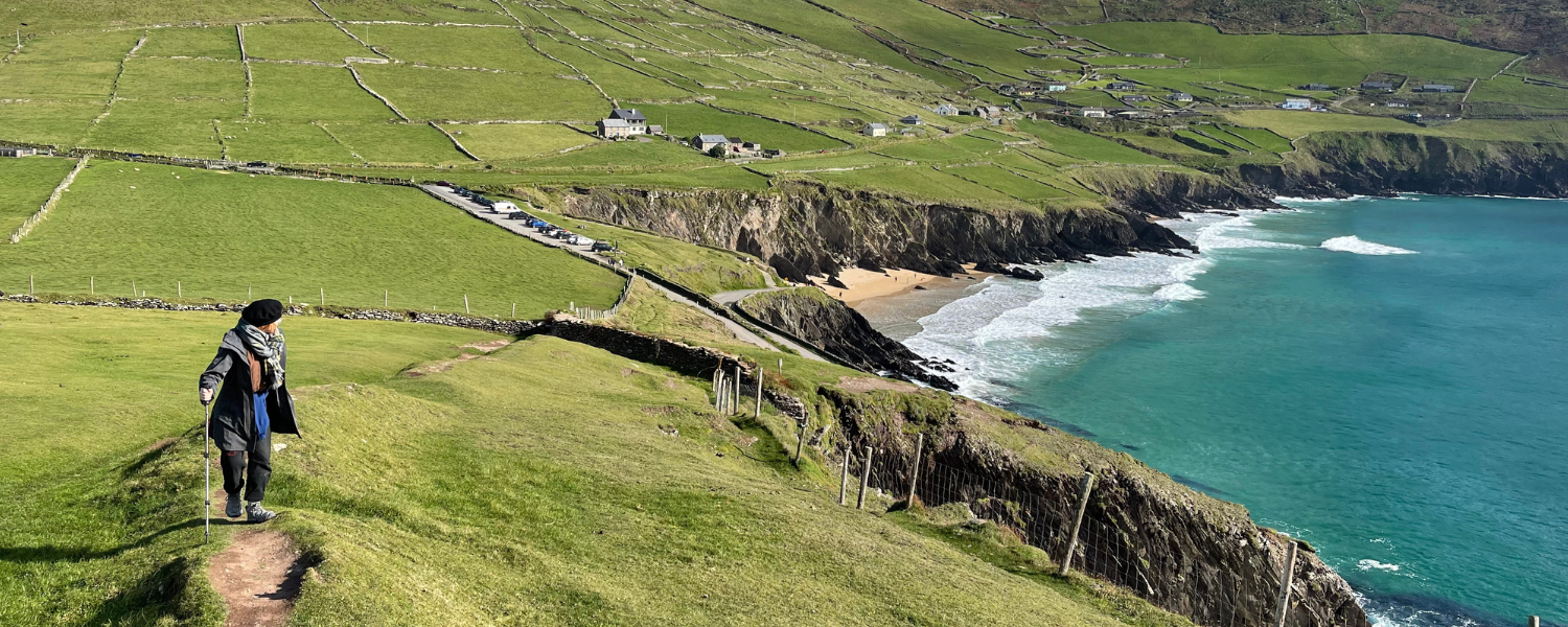 A woman wearing a coat and hat walks along a path on the top of a rocky cliff slope and looks back at a grassy green landscape and ocean below.