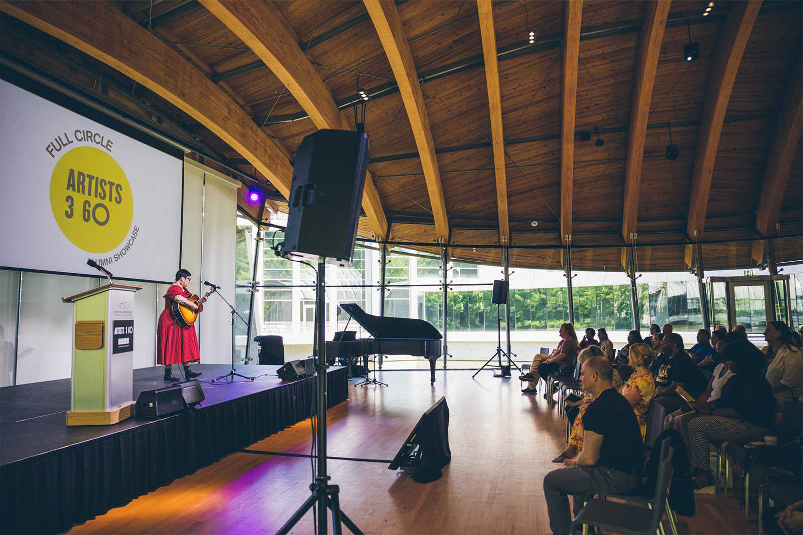 Woman in red dress stands on a stage with an acoustic guitar with the audience watching in a beautiful atrium performance hall.