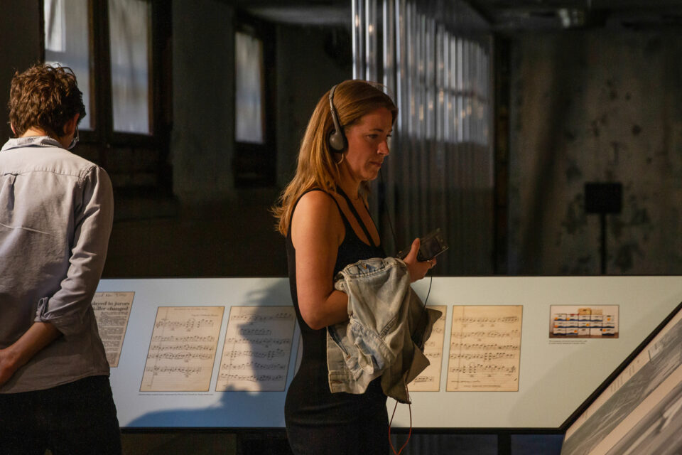 Woman stands in a jail with headphones on with the score displayed behind her.