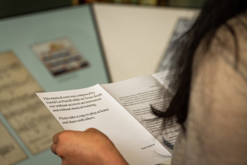 Woman holds a piece of paper that says, This musical score was composed by David Lee Powell while on death row without access to an instrument and without musical training. Please take a copy to play at home and share with others.