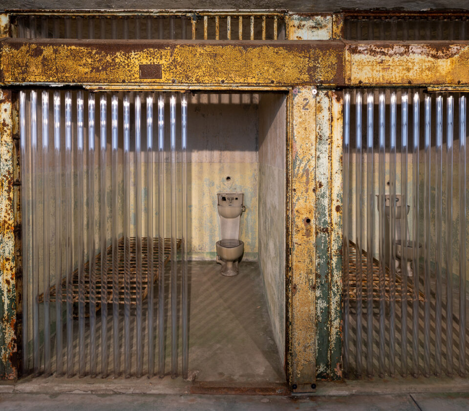 A small prison cell with bars that have been replaced with clear tubes. Paint on the walls is old, chipping, and falling off. 