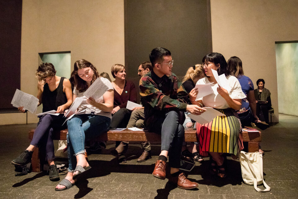 Large group of people sit, read, and exchange letters on pieces of white paper.