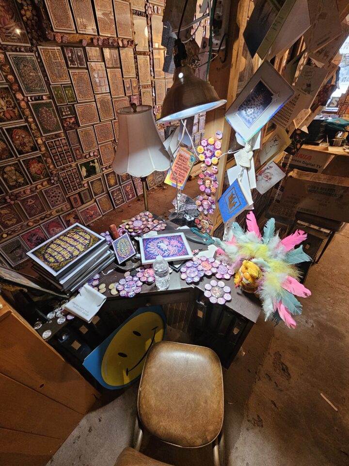 A desk with colorful materials including feathers and mixed media in a packed studio filled with art and fun trinkets.